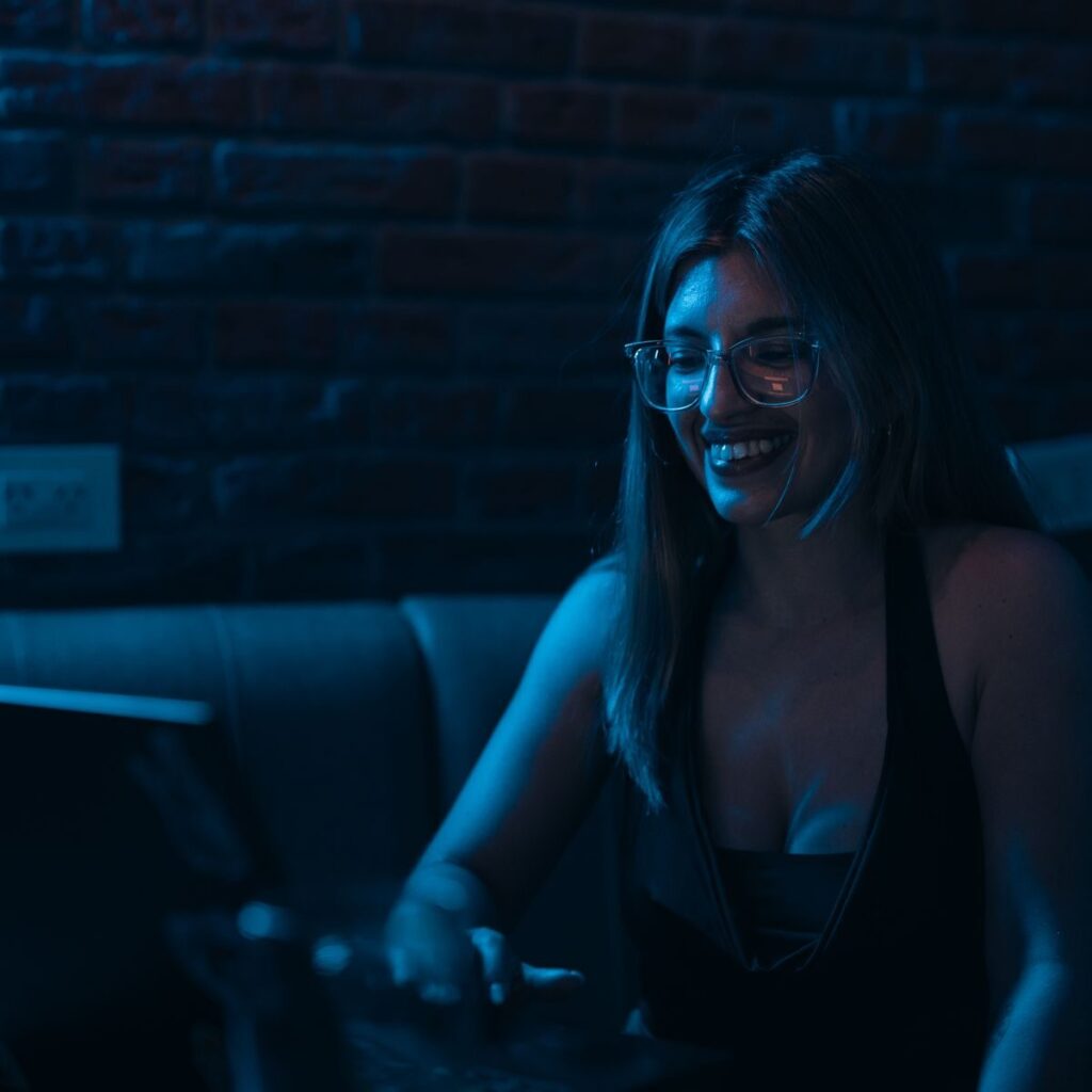 A young woman wearing glasses smiles while working on her laptop in a dimly lit room with a brick wall background.