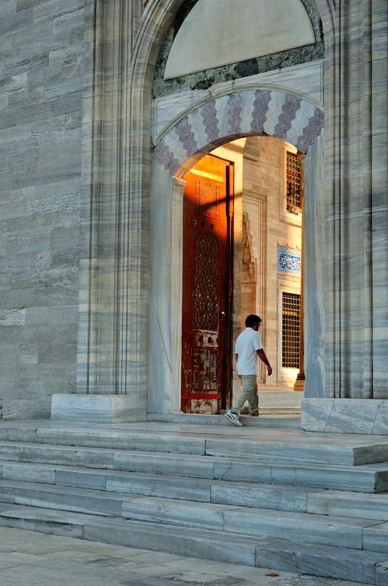 A person in a white shirt walking through a stone archway into sunlight